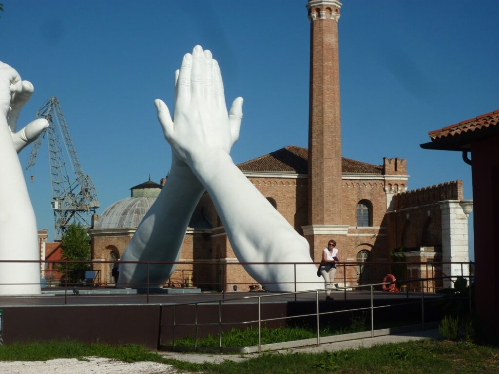 Große Skulptur zweier Arme, die sich an den Händen berühren, auf der Biennale. Im Hintergrund ein Fabrikgebude.