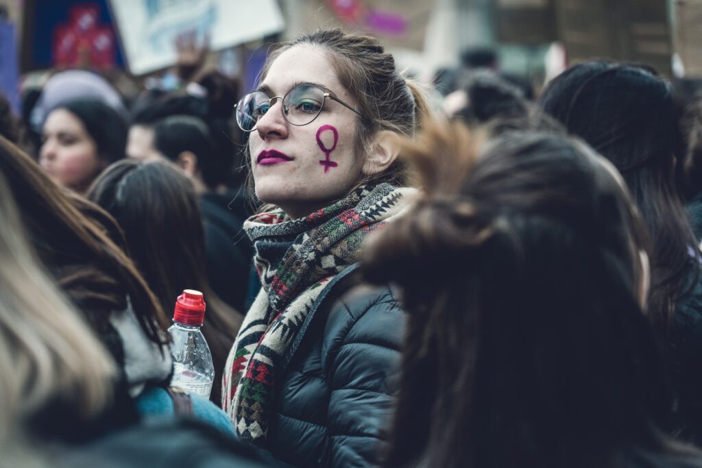 Junge Frau mit Brille auf einer Demonstration, auf der Wange hat sie den Venusspiegel aufgemalt.
