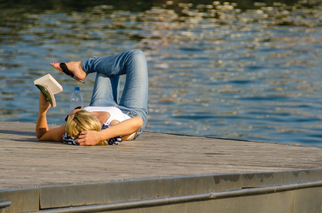Eine Frau in Sommerbekleidung mit Flip-Flops und Sonnenbrille liegt auf einem Steg am See und liest ein Buch.