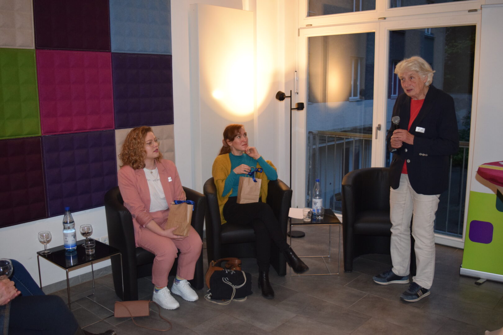 Im Foyer des Hauses Helene Weber mit großen Fenstern sitzen die Podiumsgäste Johanna Jungbluth und Nora Bossong auf Ledersesseln. Rechts davon steht die Vorsitzende des KDFB Berlin, Prof. Barbara John, mit Mikrofon in der Hand.