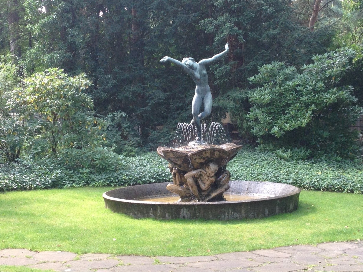 Springbrunnen in einem Garten, die Spitze des Brunnens ziert ein bronzener Frauenakt, der wie im Tanz die Arme von sich streckt.