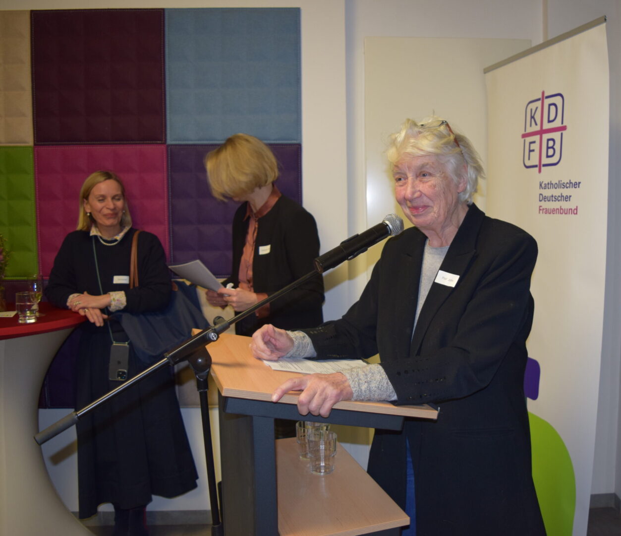 An einem Rednerpult vor dem Rollup mit dem Logo des Katholischen Deutschen Frauenbundes steht lächelnd die Vorsitzende des KDFB Berlin, Prof. Barbara John.
