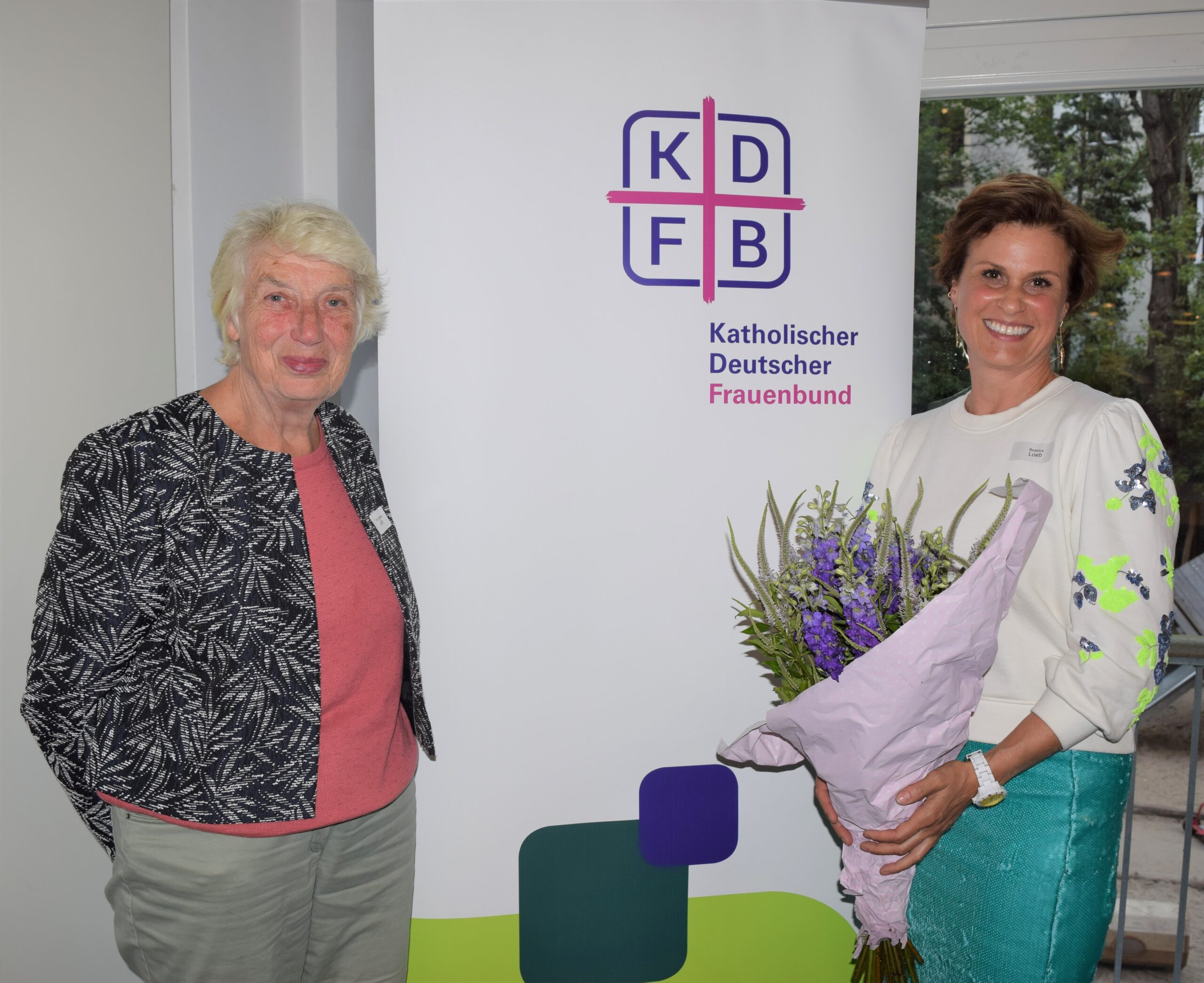 Vor dem Rollup mit dem Logo des Katholischen Deutschen Frauenbundes stehen lächelnd Prof. Barbara John, Vorsitzende des KDFB Berlin, und Festrednerin Beatrice Loeb, Vorsitzende der Gesellschaft für Christlich-jüdische Zusammenarbeit in Berlin, mit einem Blumenstrauß in der Hand.