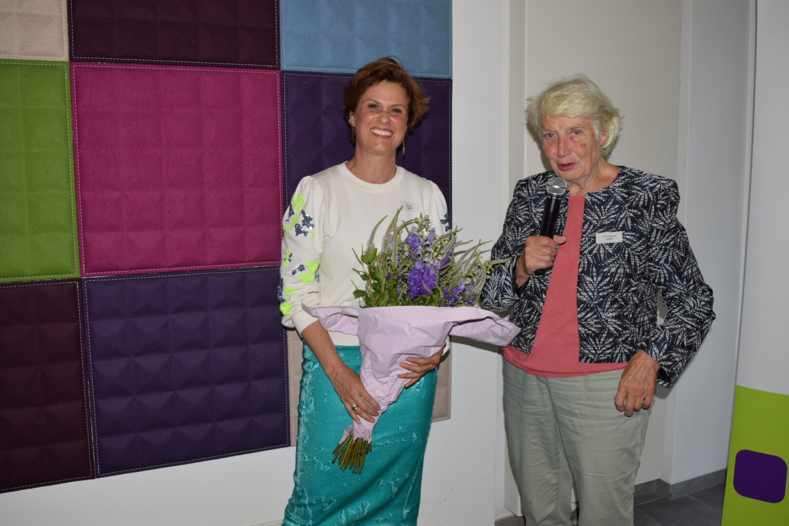 Prof. Barbara John dankt mit einem Mikrofon in der Hand der Festrednerin Beatrice Loeb, die lächelnd einen Strauß Blumen in der Hand hält.