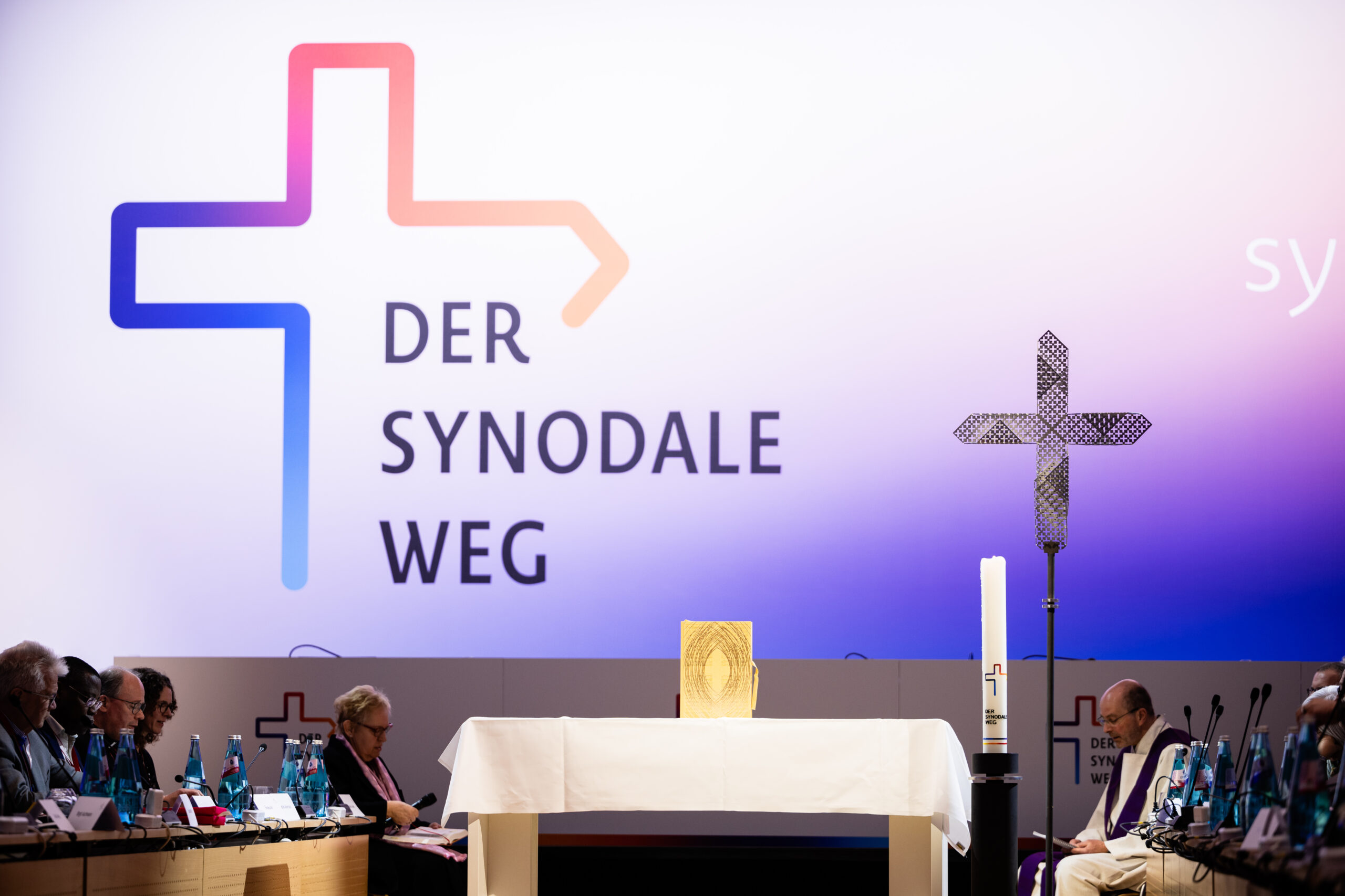 Das Foto zeigt die fünfte Synodalversammlung des Synodalen Wegs in Frankfurt: an die Wand ist das Logo des Synodalen Wegs projiziert, ddarunter sieht man ein Kreuz, eine Kerze und Teilnehmende.