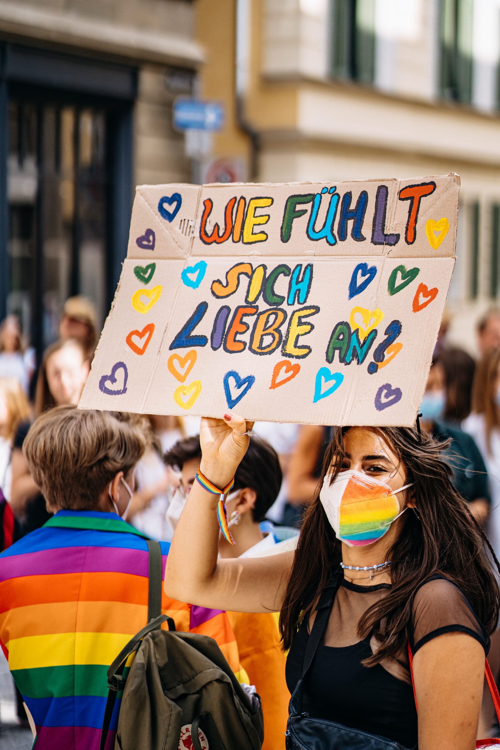 Auf einer Demo hält eine Frau mit Regenbogenarmbändchen und Regenbogen-FFP2-Maske ein Schild hoch, auf dem in bunten Buchstaben steht: Wie fühlt sich Liebe an? Daneben sind bunte Herzen gemalt.